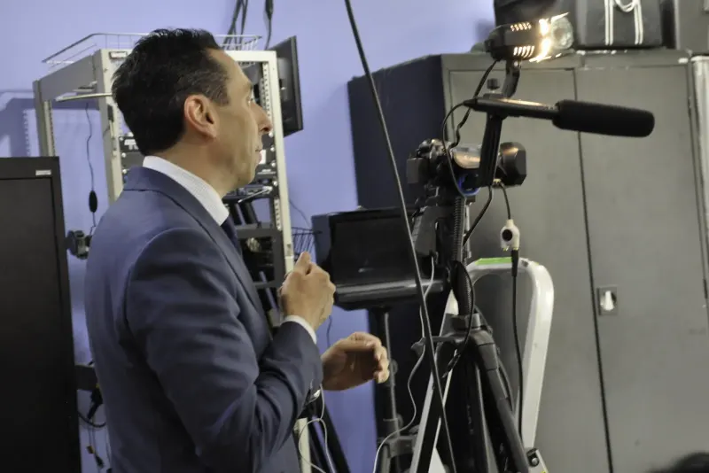 Jess in profile at a camera rig with a shotgun microphone and an equipment rack behind him