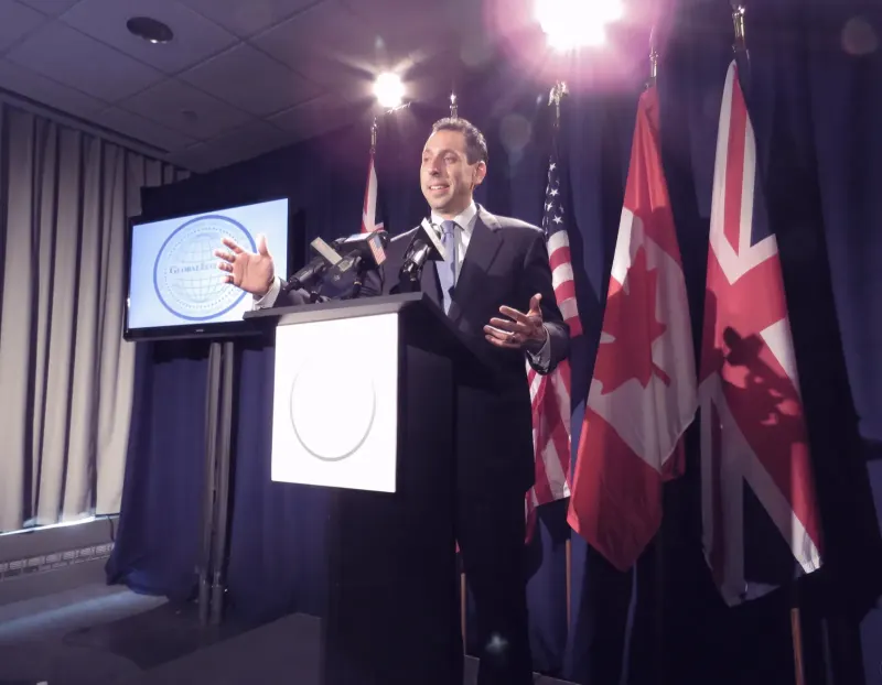 Jess at a lectern with several press microphones and flags behind him