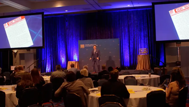 Jess on a ballroom stage in front of blue curtains, audience tables in the foreground